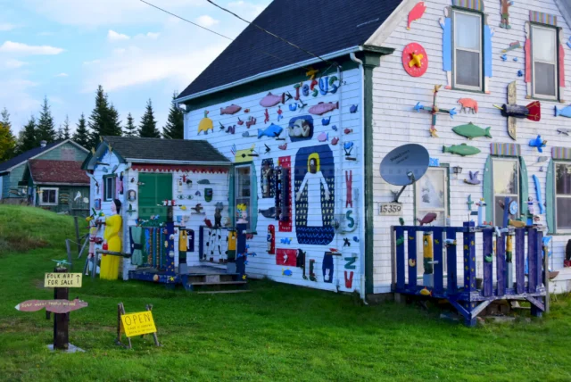 The exterior of the house is full of primitive art and the sale sign points to the back.