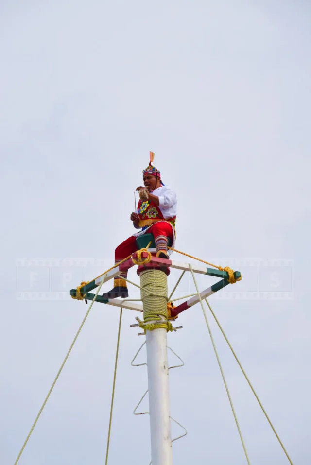 native mexican sitting on top of the pole and playing the flute for four dancers.