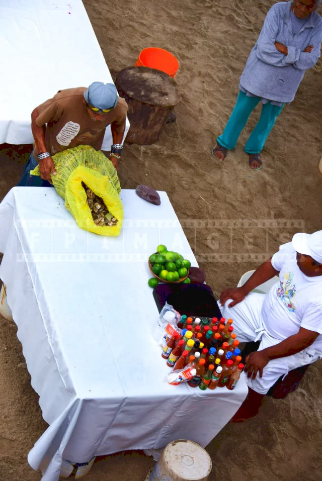 Fisherman delivering a bag of freshly caught oysters to a beachfront vendor, the table is set with limes and condiments